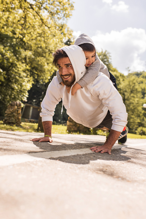 Young smiling father holding his son on back and exercising push-ups in the park.